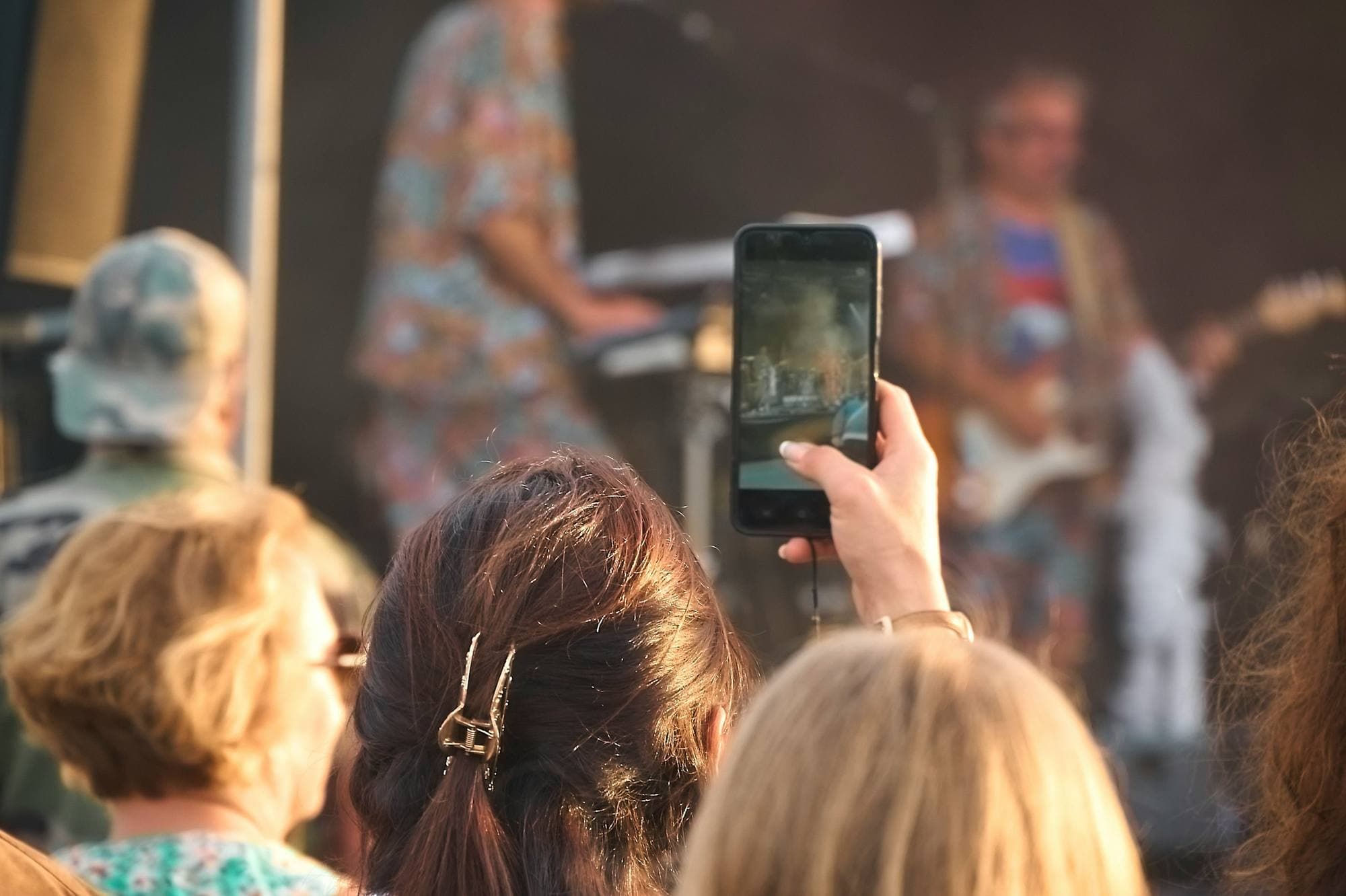Une spectatrice lève son téléphone pour filmer un groupe qui joue en plein air, en journée