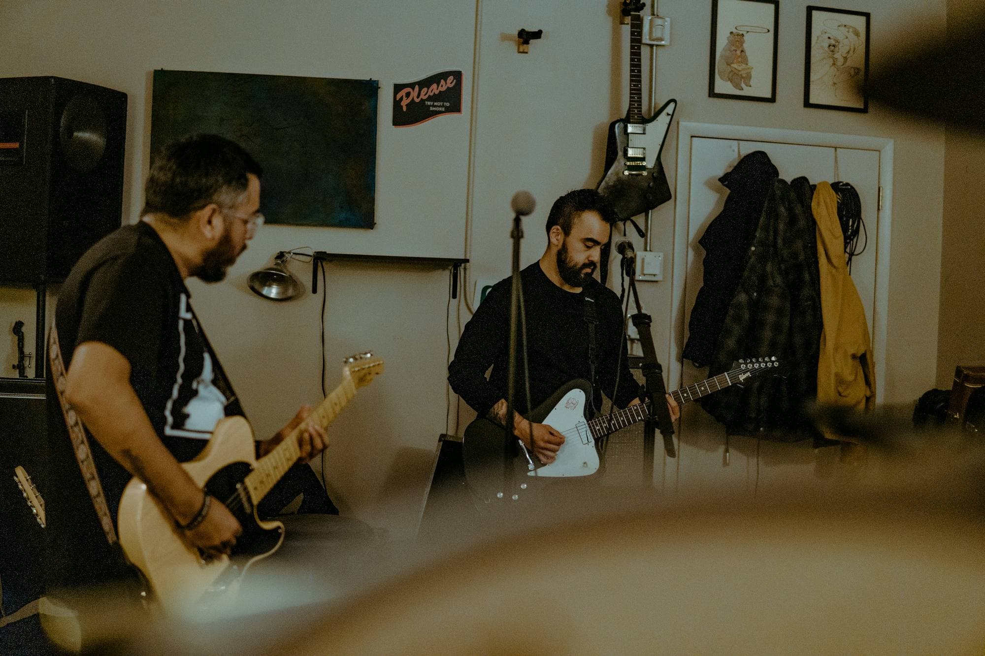 Deux guitaristes jouent en formation intime dans une petite pièce, manteaux et cadres au mur