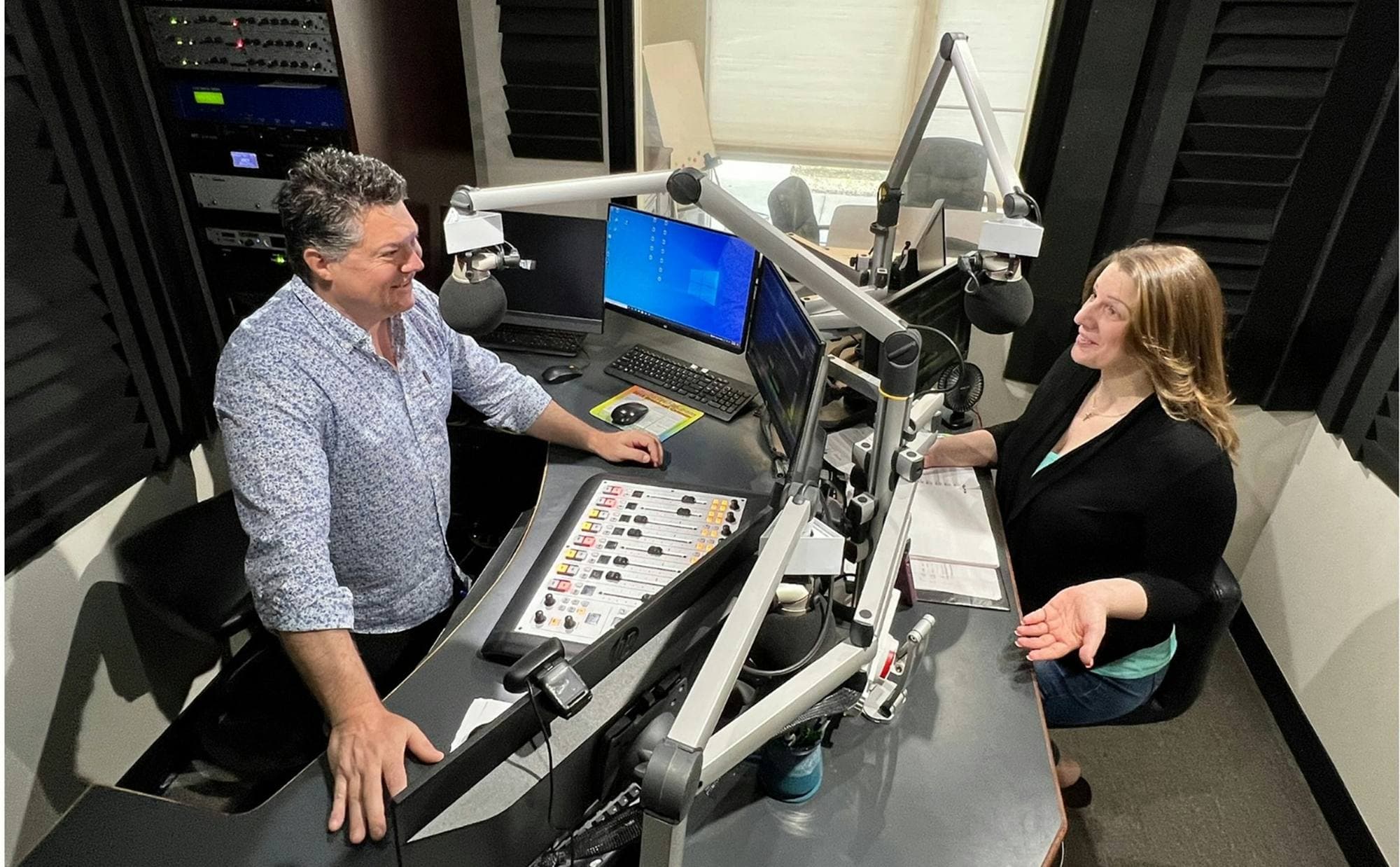 Deux animateurs discutent à la console d'un studio radio, micros sur bras articulés, table de mixage et écrans
