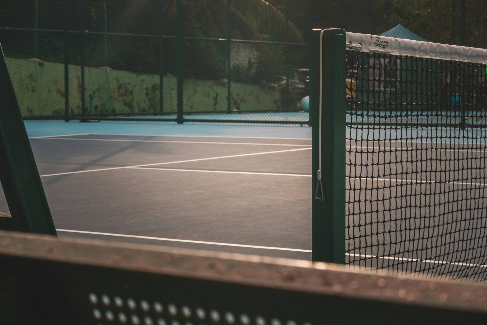 Court de tennis extérieur vu derrière le poteau du filet, bordé d'une clôture grillagée